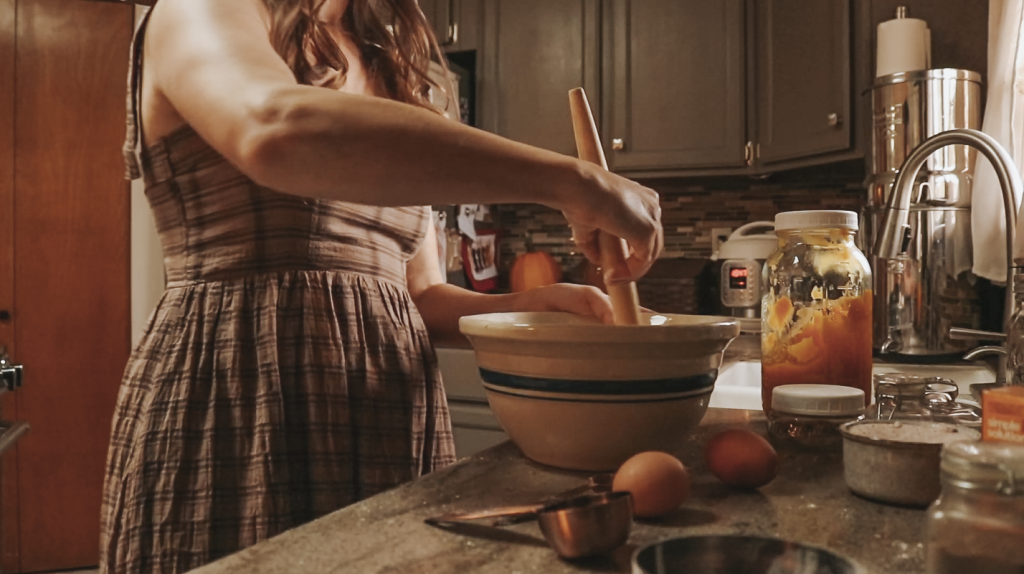 Christian homemaker wearing a brown plaid dress is standing at her kitchen counter and mixing food in a large ceramic bowl.