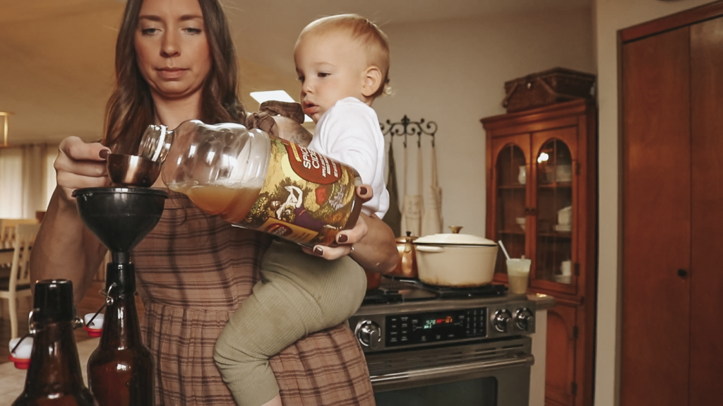 Christian homemaker wearing a brown plaid dress and carrying her young toddler on her hip pours some apple cider through a funnel into a brown glass bottle.