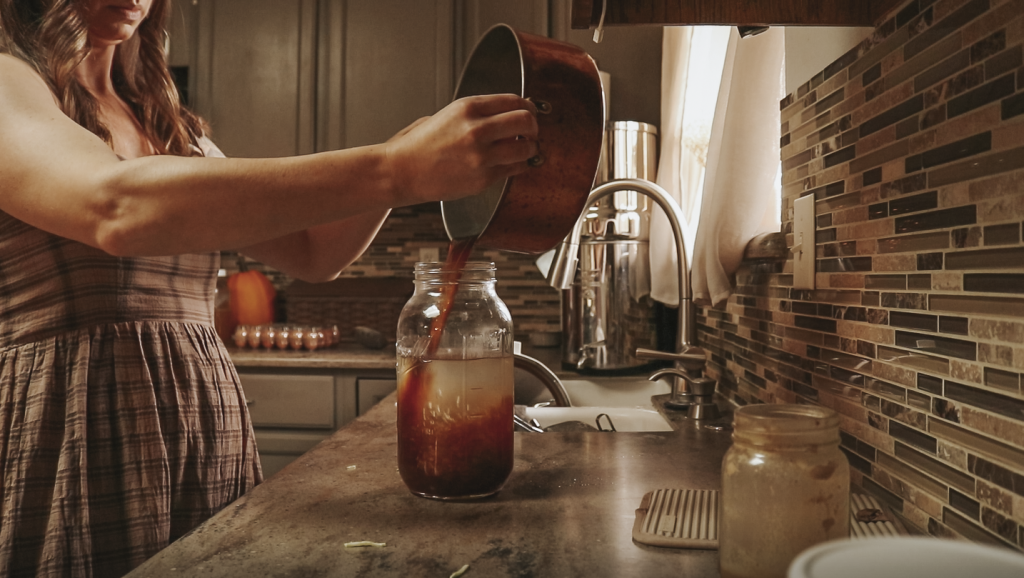 Christian homemaker wearing a brown plaid dress pours a pot homemade broth into a gallon size glass mason jar.