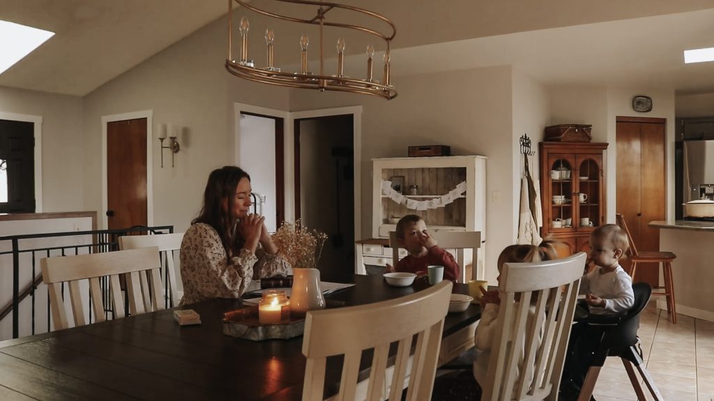 Christian homemaker and mother prays while siting with her three children at the dining room table.