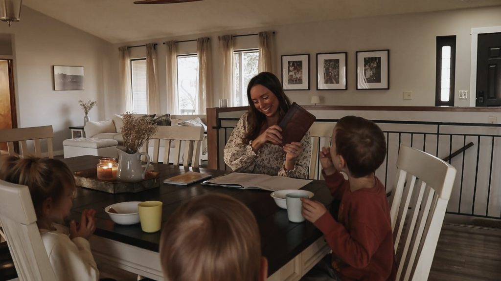 Homemaker and mother sits with children at dining room table while smiling and reading to her children.
