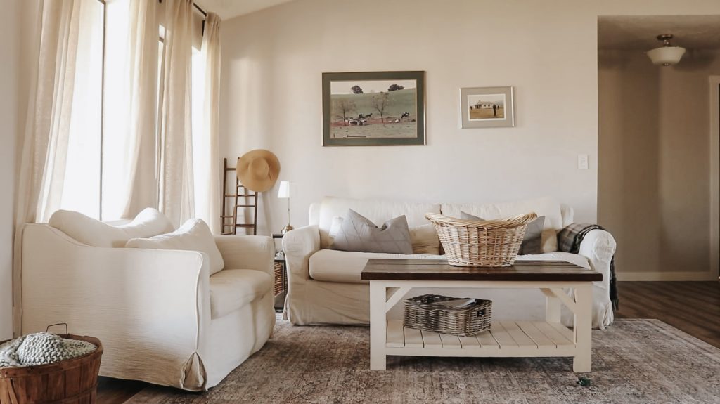 Bright and airy living room with white couch and chair and wicker laundry basket sitting on a wooden coffee table.