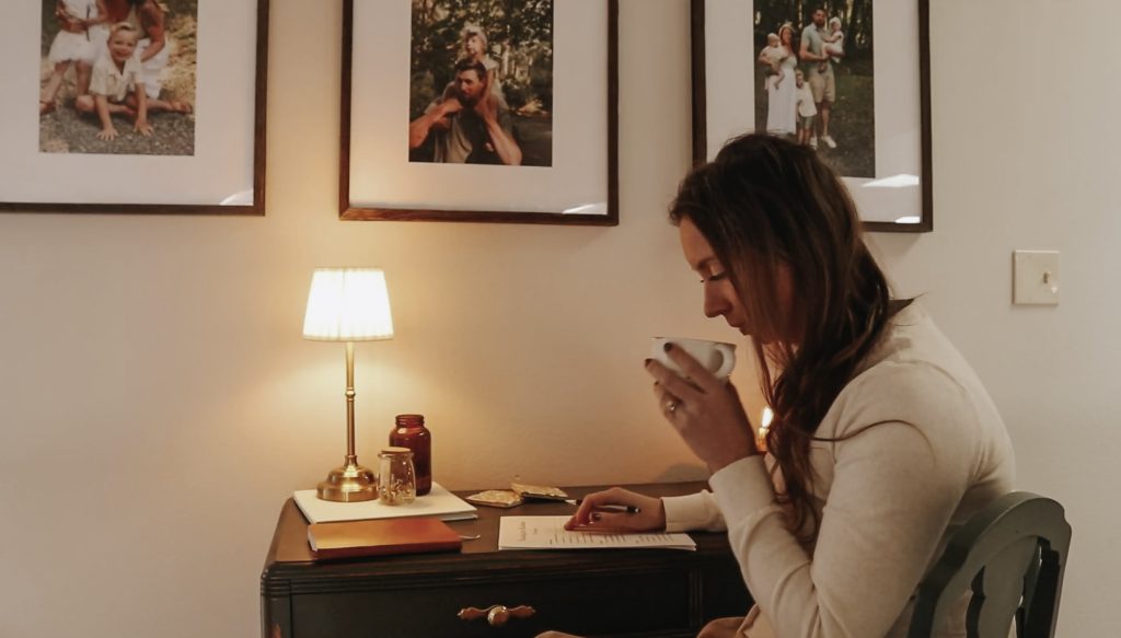 Homemaker sips coffee while writing and sitting at a desk lit by a lamp.