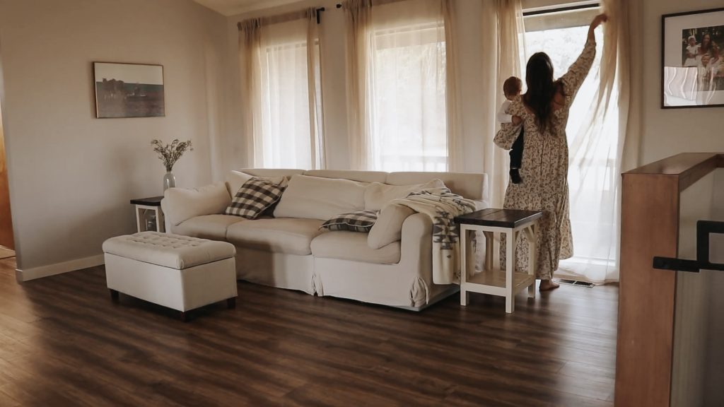 Homemaker opens light and airy curtains in living room while holding baby on her hip. The living room is tidy and ordered, ready for the day ahead.
