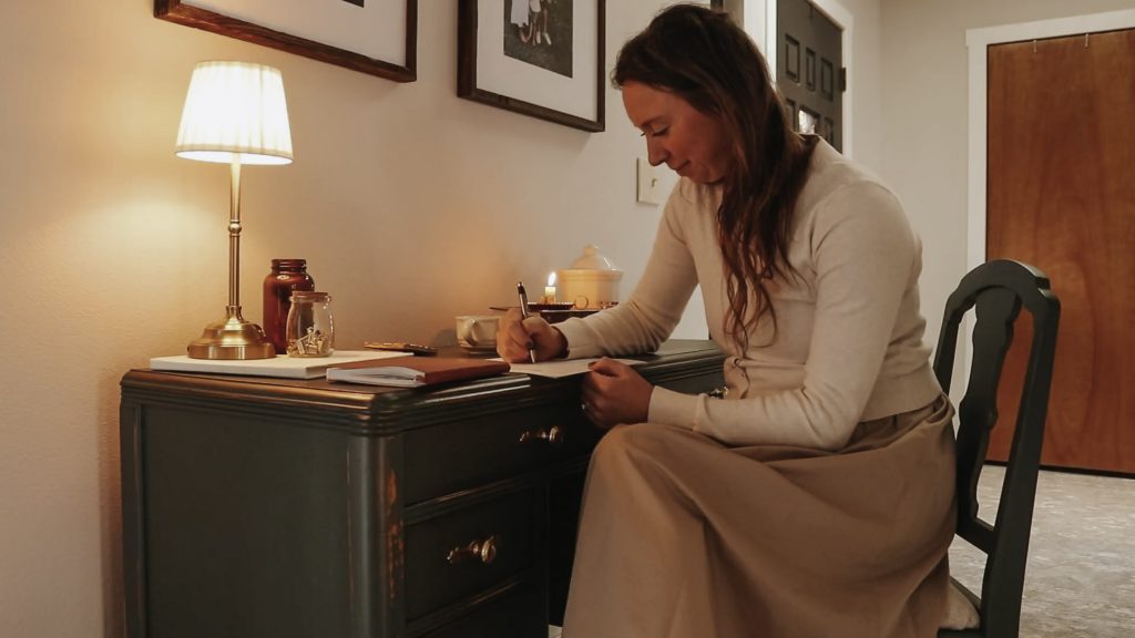 Homemaker sits at desk with long dress and cardigan writing in a notebook.