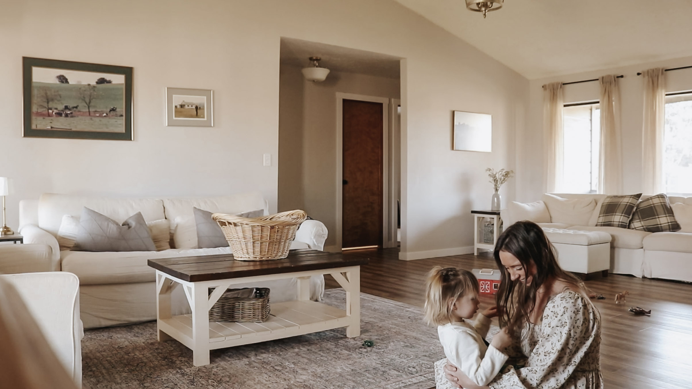 Homemaker and mother sits on the floor of the living room with her toddler and smiles while holding her in her lap.