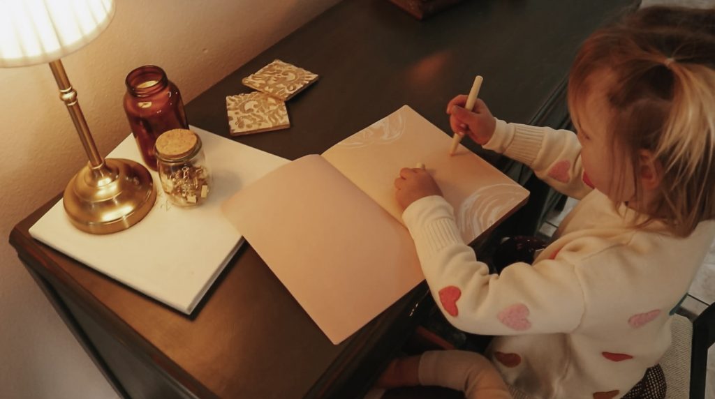 Toddler child sits at desk lit by a table lamp and colors in a notebook.