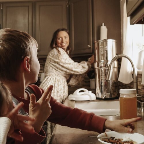 Christian homemaker and mother looks over her children while preparing food in the kitchen while wearing a long, flowing dress.