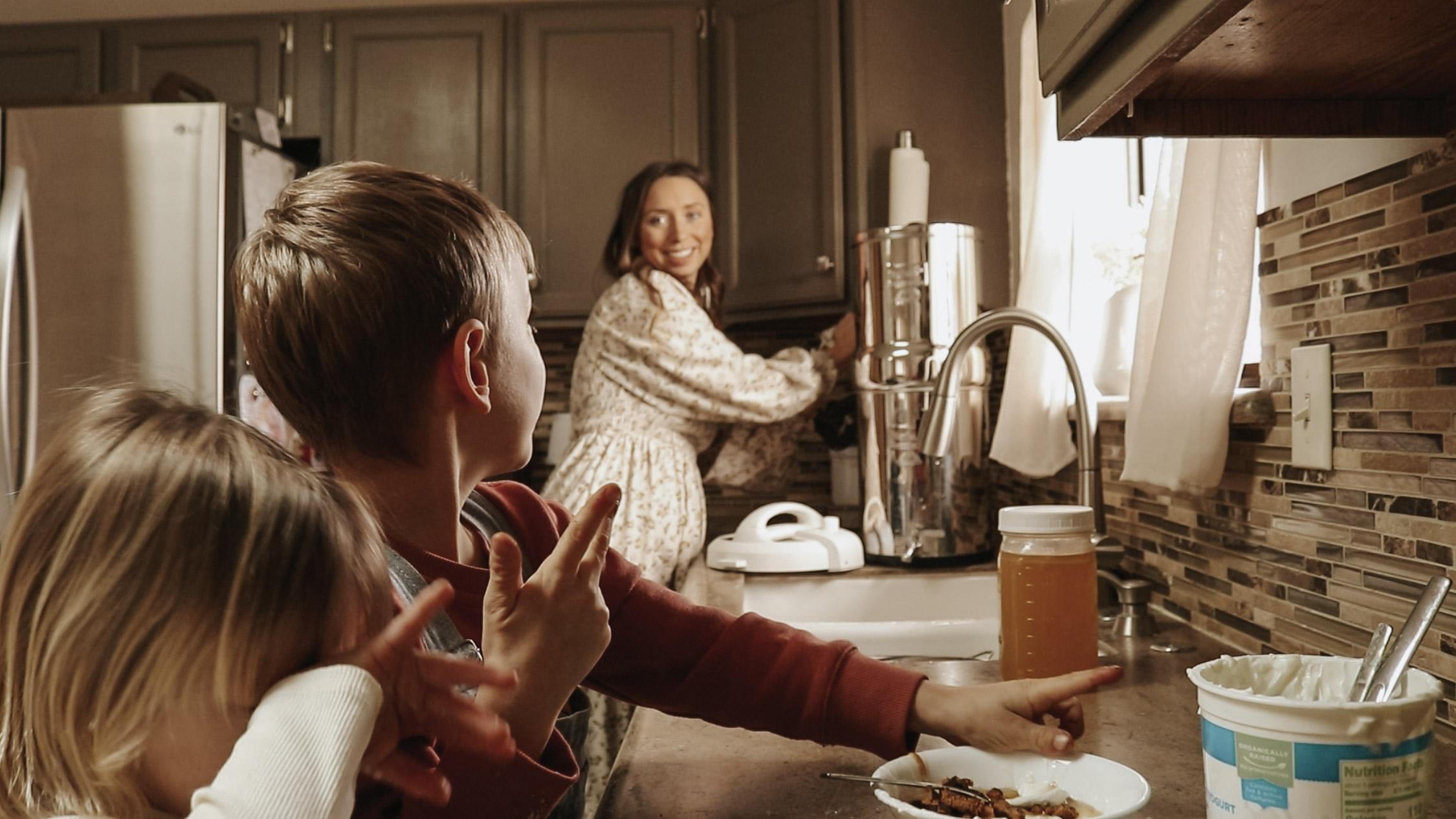 Christian homemaker and mother looks over her children while preparing food in the kitchen while wearing a long, flowing dress.