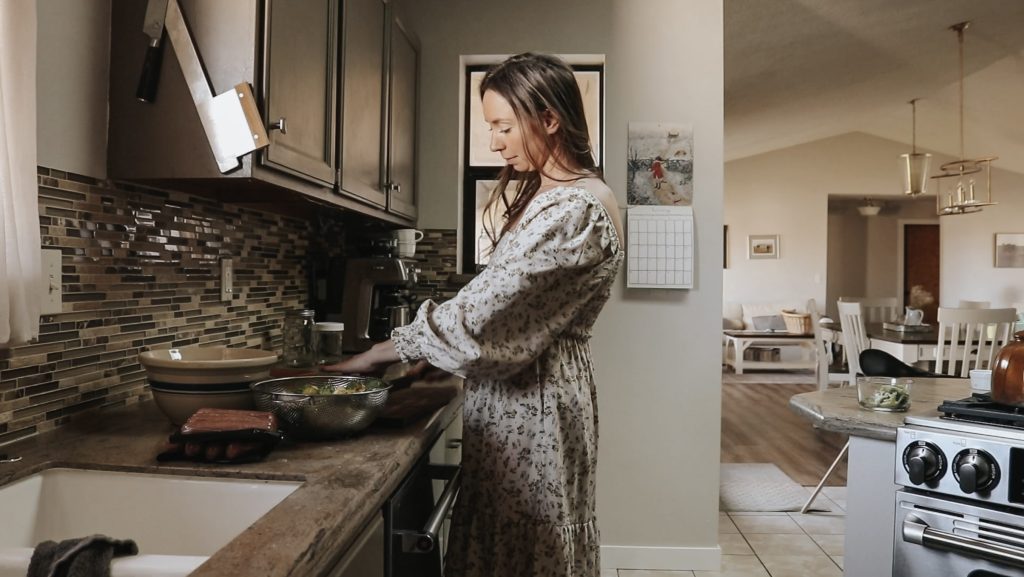 Homemaker prepares a meal at the kitchen counter while wearing a long, floral dress.