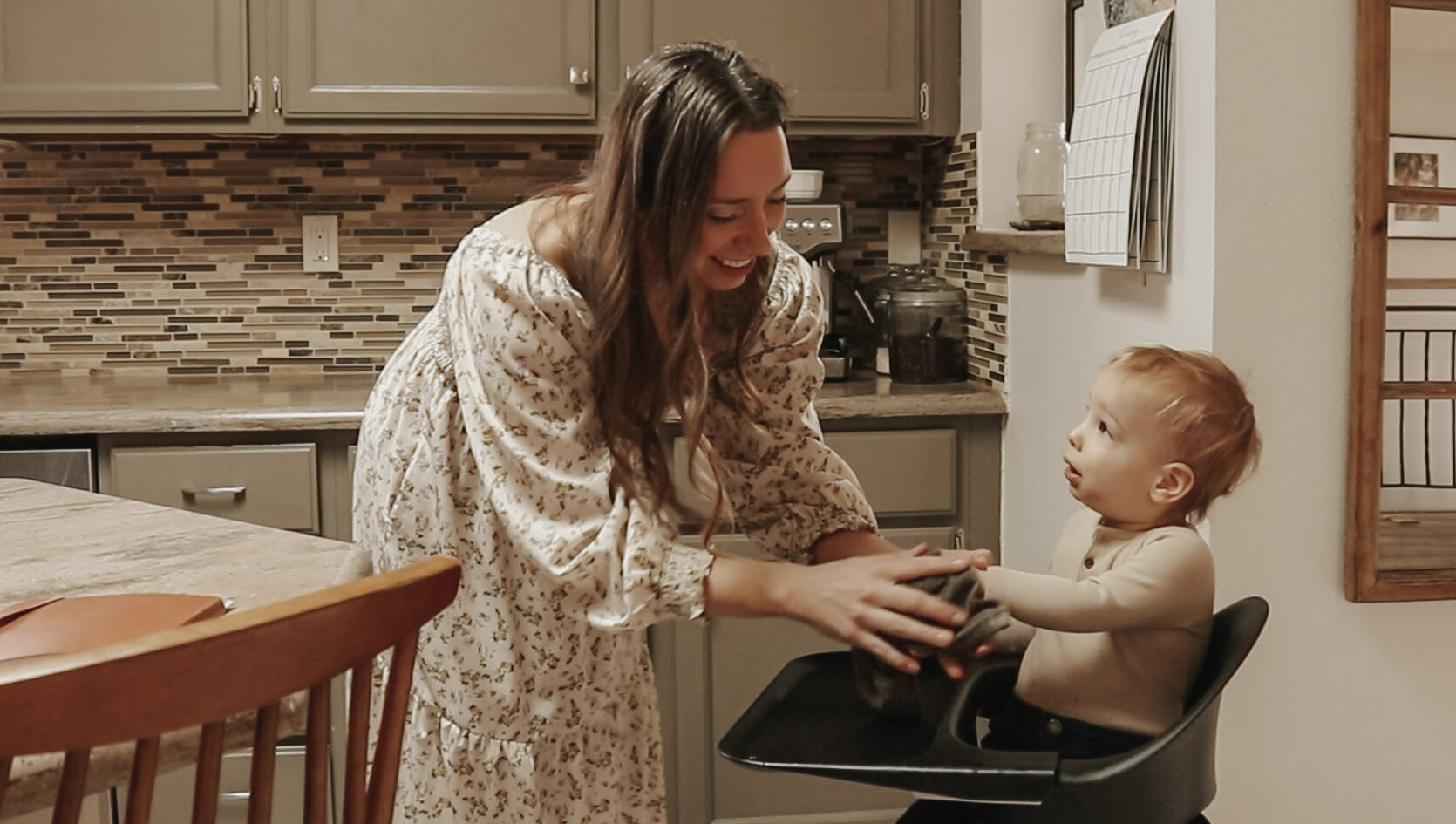 Homemaker and mother wearing a floral dress cleans baby sitting in high chair after meal.