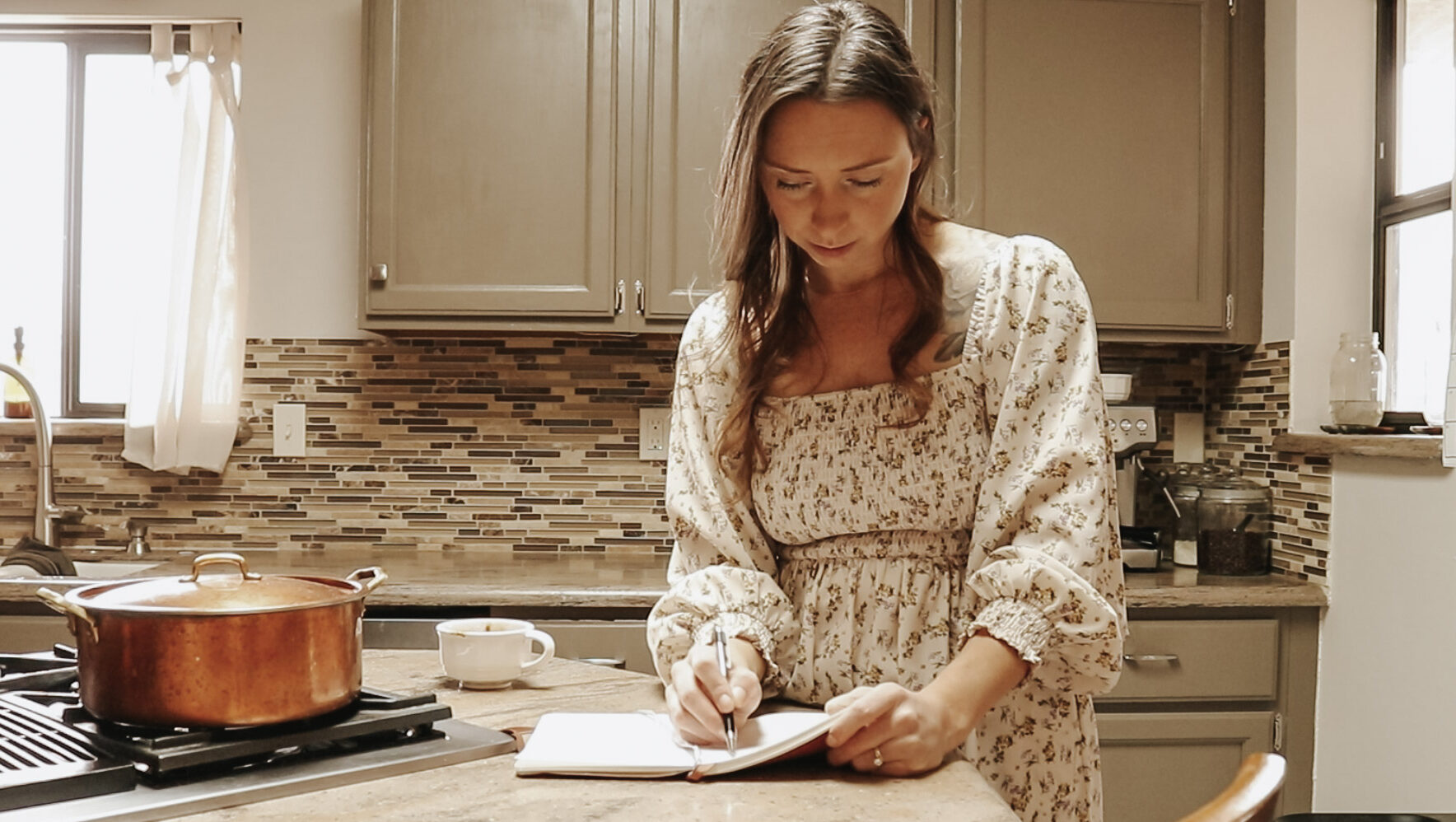 Homemaker stands at kitchen counter and writes in a notebook wearing a long, floral dress.