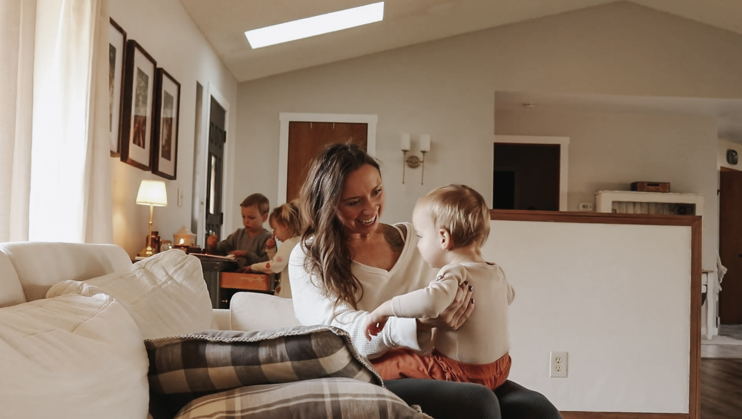 Homemaker and mother smiles while holding her baby on a white couch with her children in the background.