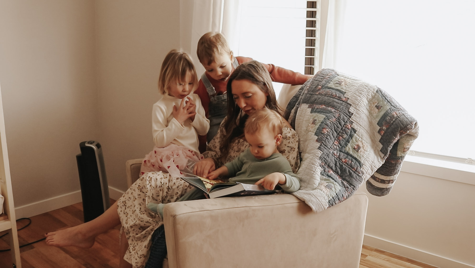 Homemaker and mother sits in a beige rocking chair with her children while reading a book.