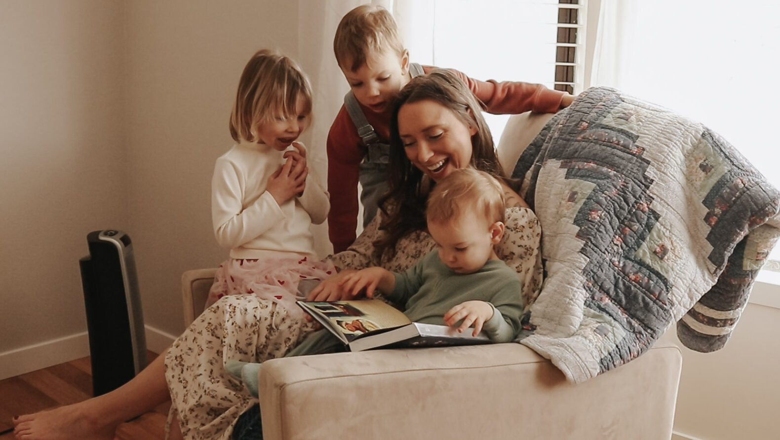 Homemaker and mother sits in a beige rocking chair with her children while reading a book.