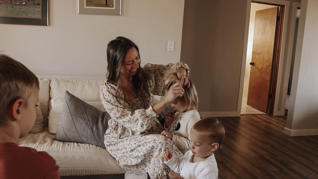 Homemaker and mother in long, floral dress smiles while fixing her daughter's hair.