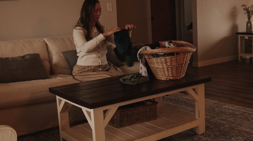 Homemaker sits on a white couch and folds laundry out of a wicker basket.