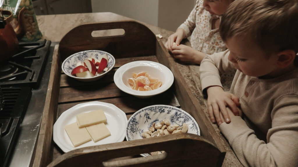 Children sit at a wooden tray with little plates of snacks.