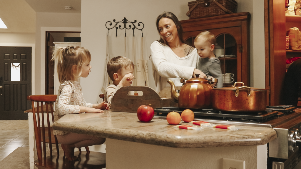 Christian homemaker stands in her kitchen holding her baby and her two older children are getting a lunch ready and placed on a tray.