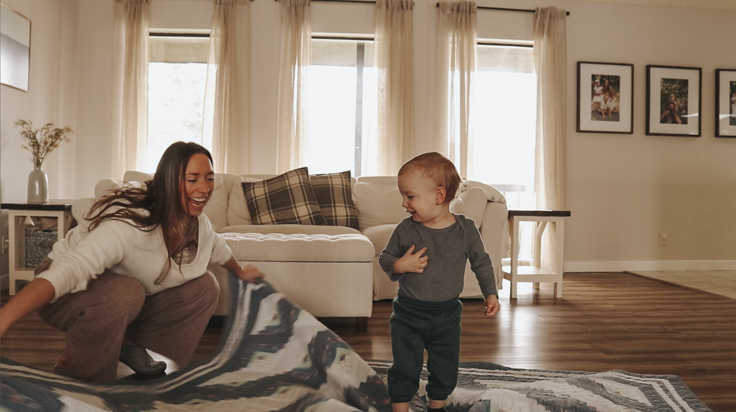 Christian homemaker is trying to spread a blanket on her living room floor, but her toddler is walking on top of it and she is laughing!
