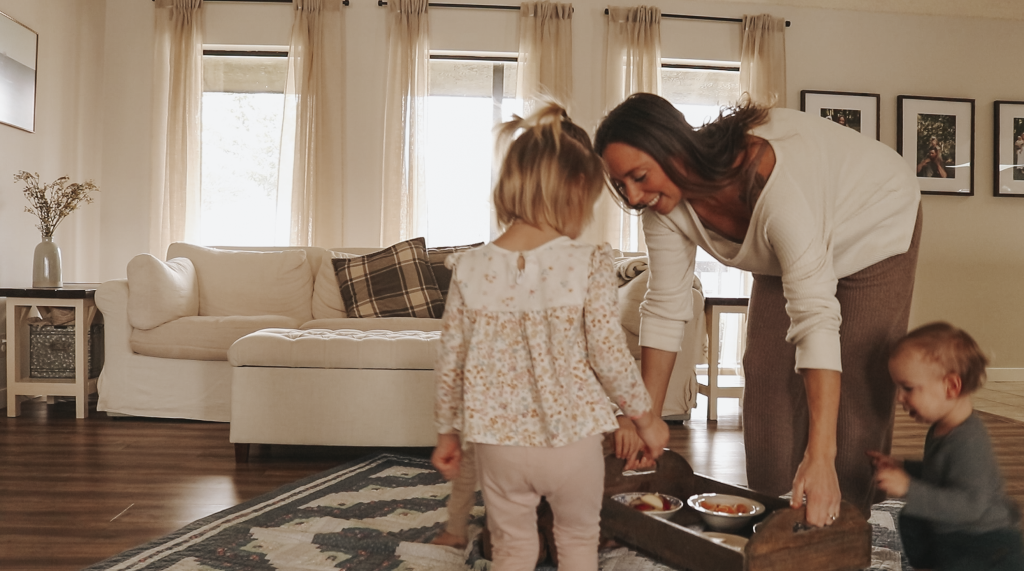 Christian homemaker wearing a cream colored top and brown pants places a tray with snacks on top of a blanket on the floor.
