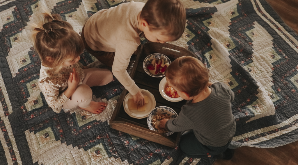 Three children reach for and eat snacks off a tray on top of a blanket on the living room floor.