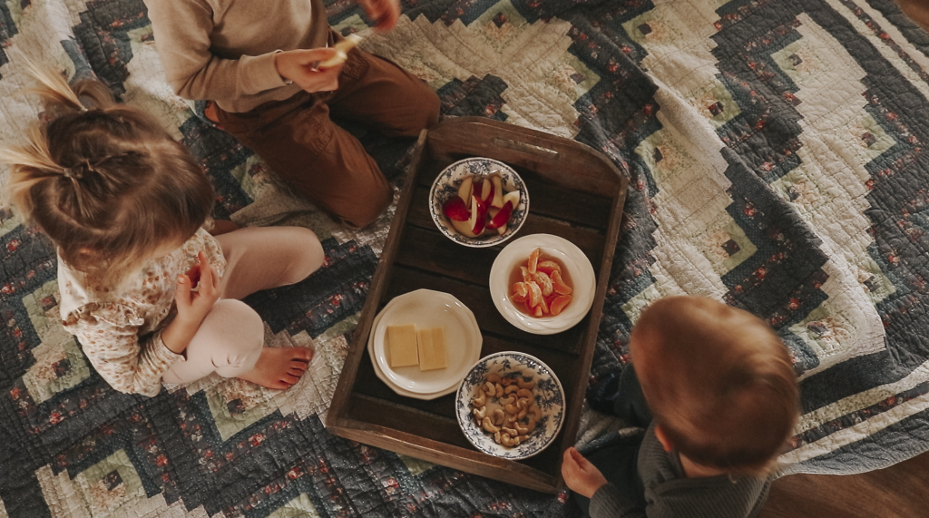 Three children sit around a tray of snacks on top of a blanket on the floor in the living room.