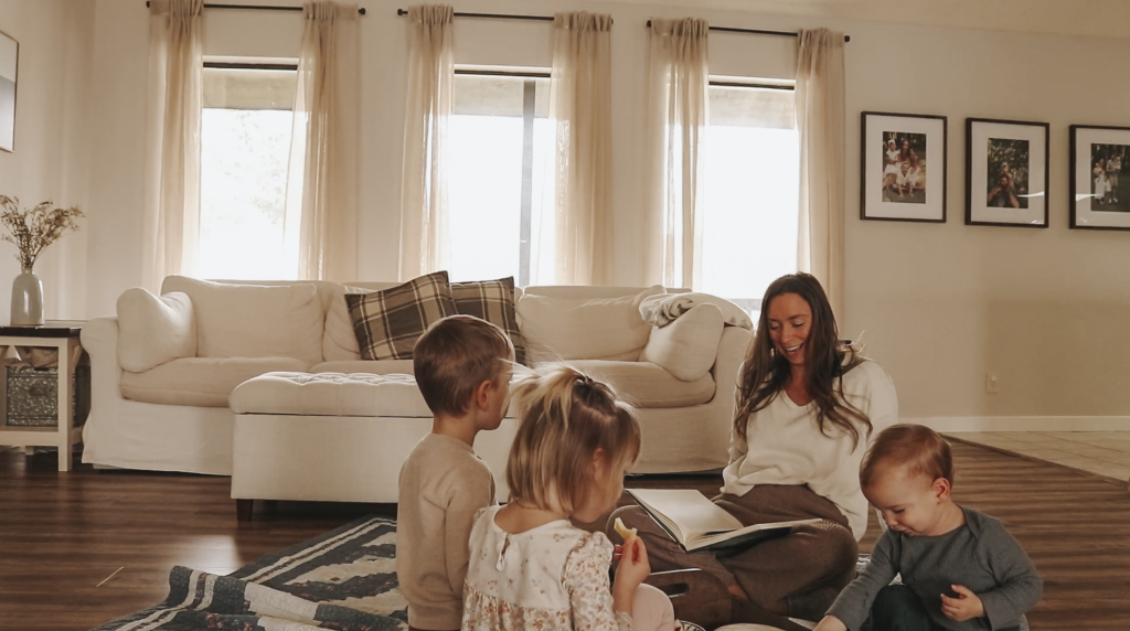 Christian homemaker sits  on a blanket on the floor with her children and reads them a book while they eat snacks off of a tray.