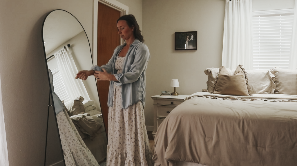 Christian homemaker wearing a white floral dress and a button up denim shirt is standing in front of her long mirror in her bedroom after getting dressed for the day.