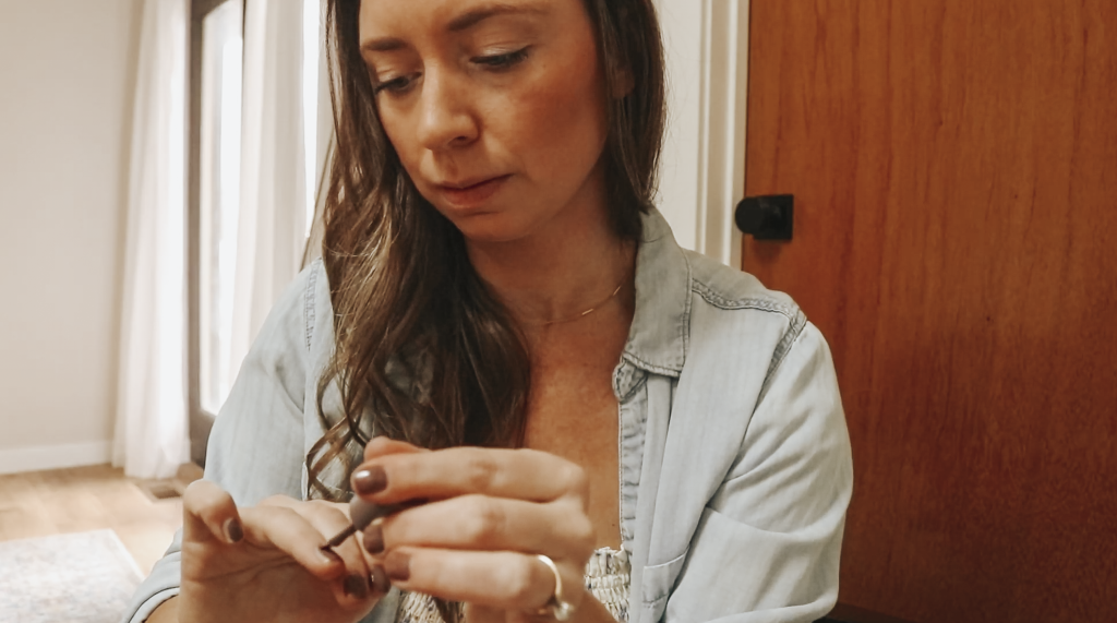 Close up of a Christian homemaker wearing a denim button up shirt sitting at her nightstand in her bedroom and painting her nails a brown color.