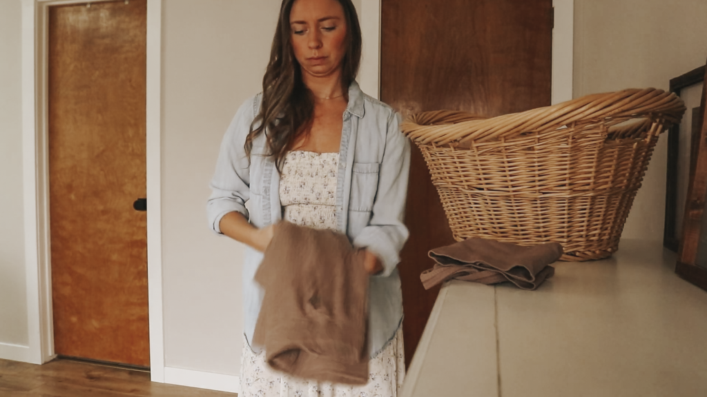 Christian homemaker wearing a white floral dress and a blue denim button up shirt is standing next to a basket of laundry and is carrying a brown apron in her arms.