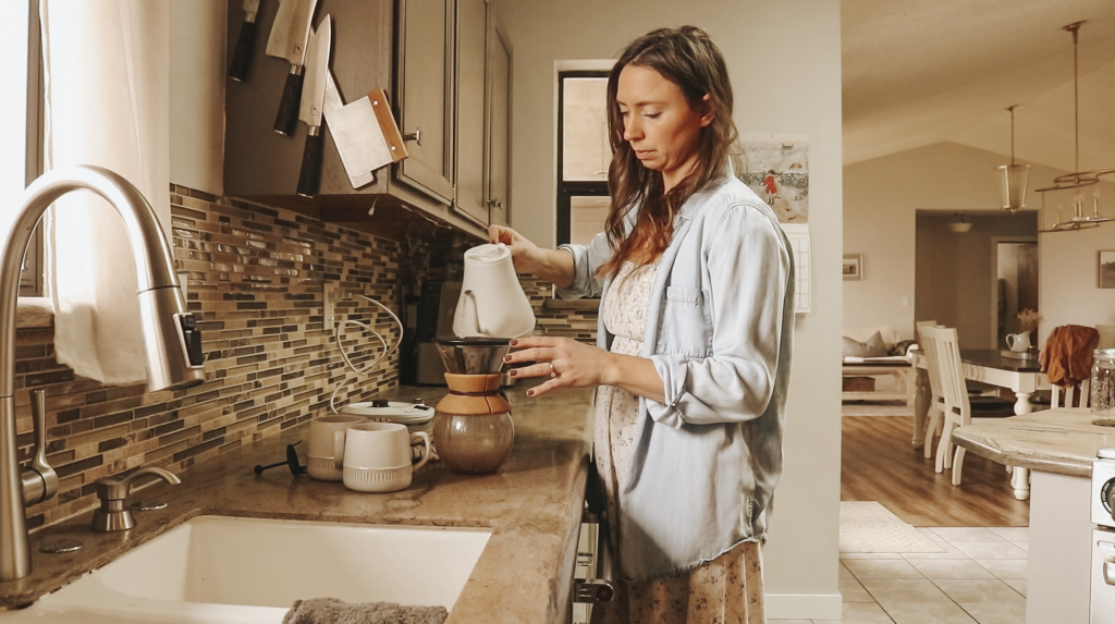 Christian homemaker wearing a white floral dress and a blue denim button up shirt is standing next to the kitchen sink pouring a water pitcher into a coffee maker.