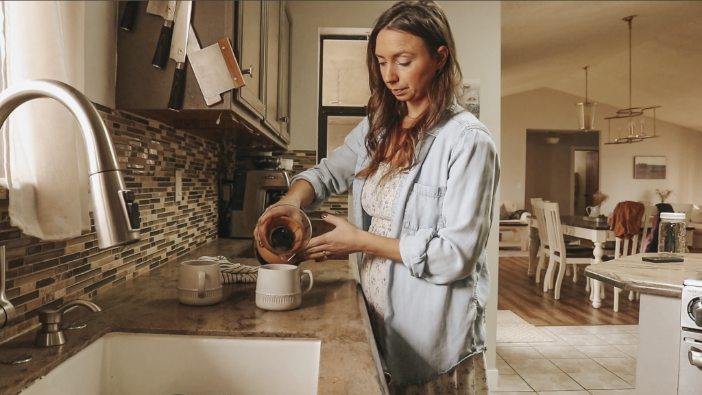 Christian homemaker wearing a white floral dress and a blue denim button up shirt is standing next to the kitchen sink pouring coffee into a mug.