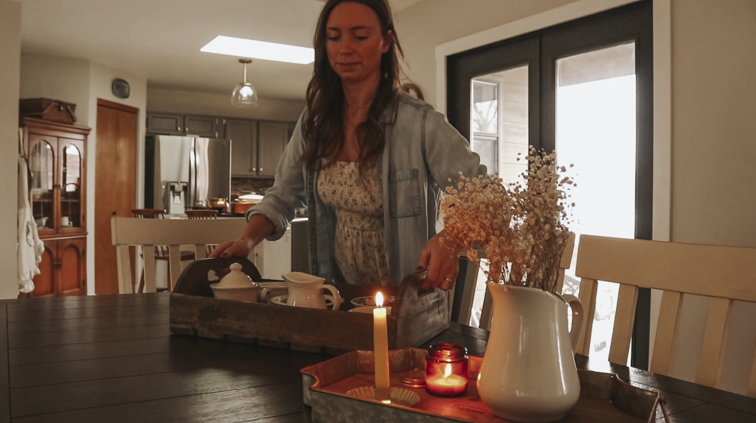 Christian homemaker wearing a white floral dress and denim button up shirt places a tray with oatmeal toppings on the dining room table.