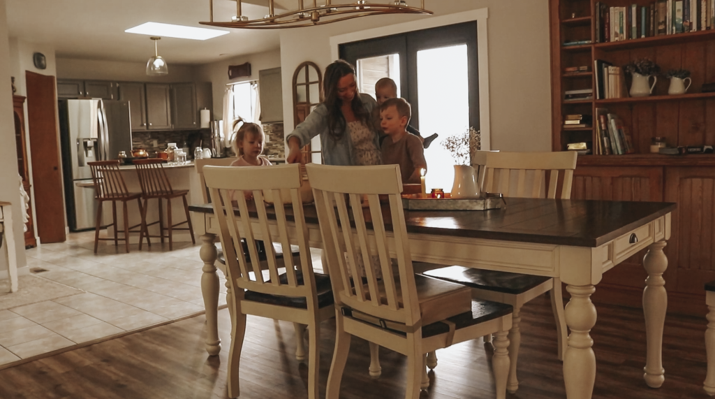 A farther away view of the Christian homemaker and her three children standing at the kitchen table spread of cooked oatmeal and toppings.