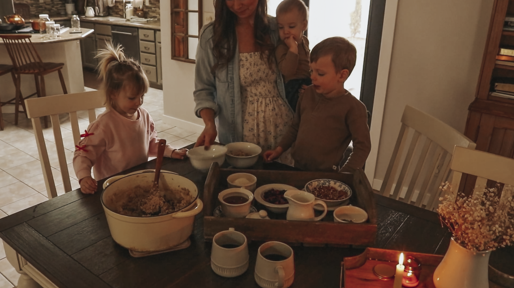 Christian homemaker wearing a white floral dress and button up denim shirt is standing at the kitchen table with her two older children and a baby on her hip. On the table there is a tray with oatmal toppings, a pot of cooked oatmeal and some bowls.
