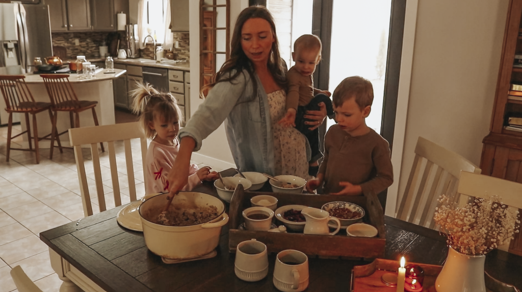 Christian homemaker wearing a white floral dress and a denim button up shirt stands at the kitchen table with her three children and is taking some cooked oatmeal from a pot to put in a smaller bowl.