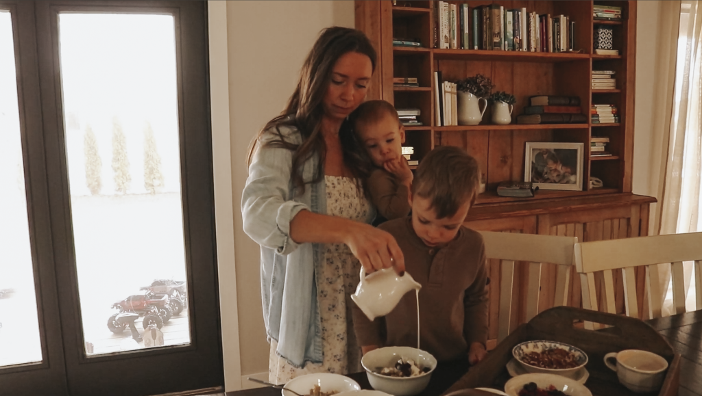 Christian homemaker wearing a white floral dress and button up denim shirt stands behind her oldest son with a baby on her hip and is pouring some cream from a white glass pitcher onto his hot bowl of oatmeal.