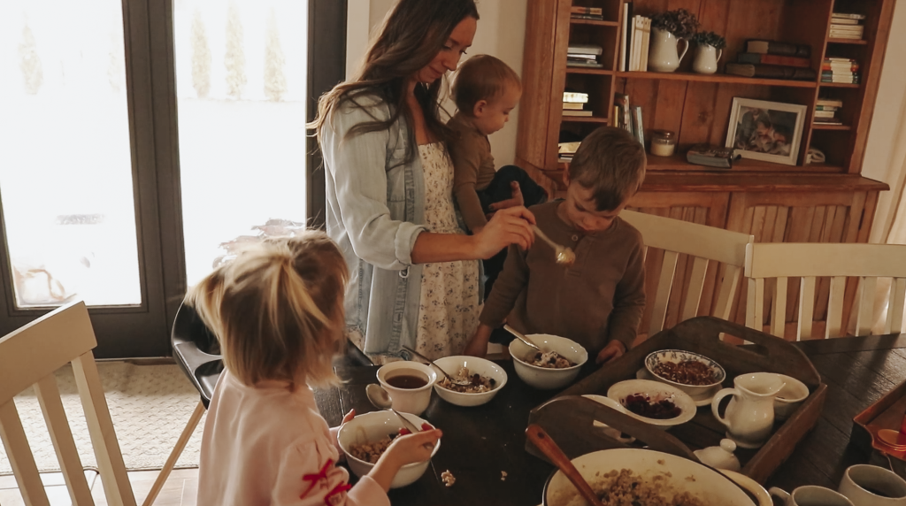 Christian homemaker wearing a white floral dress and button up denim shirt stands behind her oldest son with a baby on her hip and is helping her elementary aged sone with his hot bowl of oatmeal.