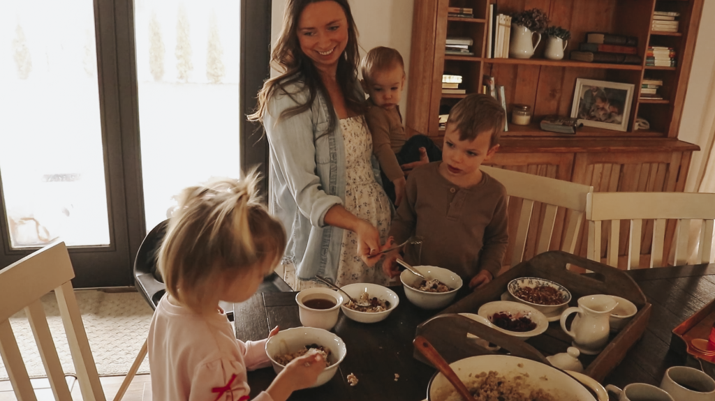 Christian homemaker wearing a white floral dress and denim button up shirt is smiling at her young daughter as she gets ready to take a bite of her oatmeal with various toppings.