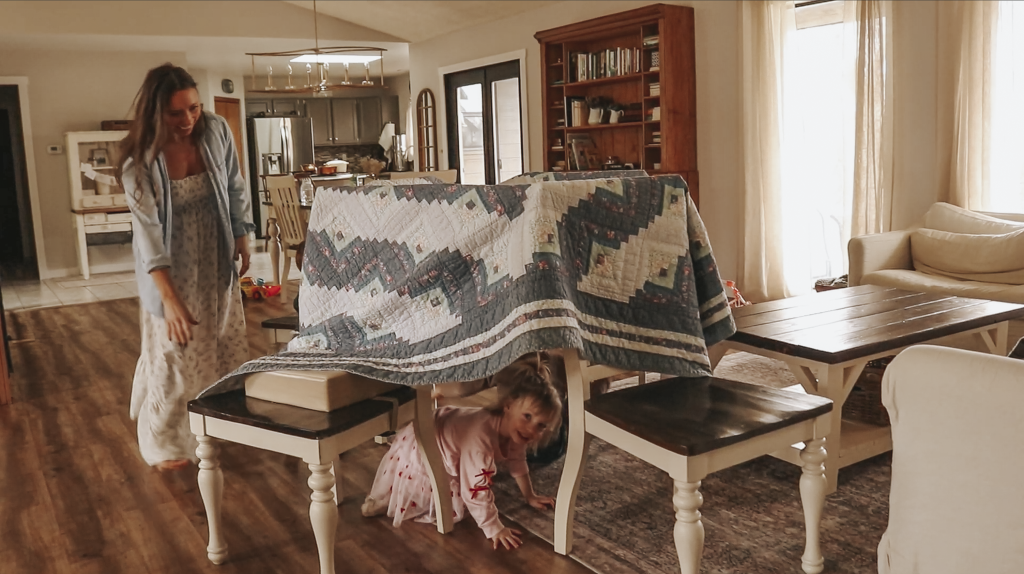 Toddler girl plays under a blanket fort made between two wooden chairs her Christian mother stands beside watching and smiling.