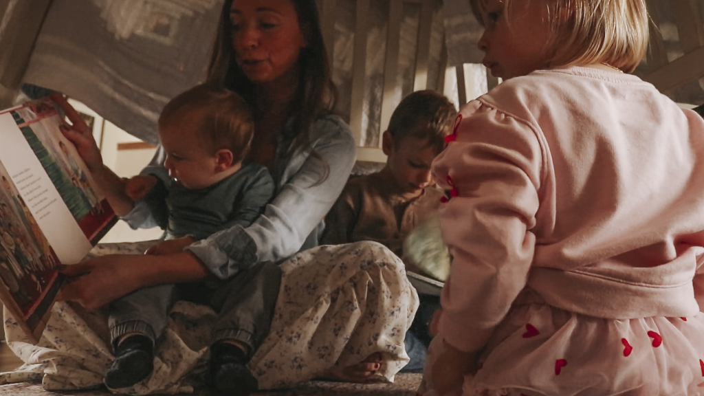 Christian mom reads to her 3 children while sitting under a blanket fort. Her toddler is sitting in her lap.