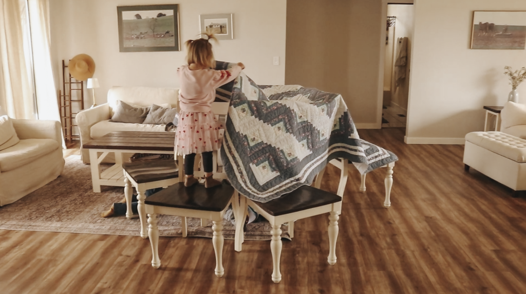 Toddler girl plays on a blanket fort made between two wooden chairs.
