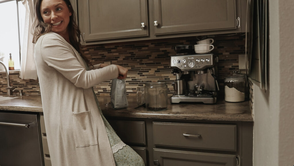 Christian homemaker smiles while making coffee and wearing a long, floral dress.