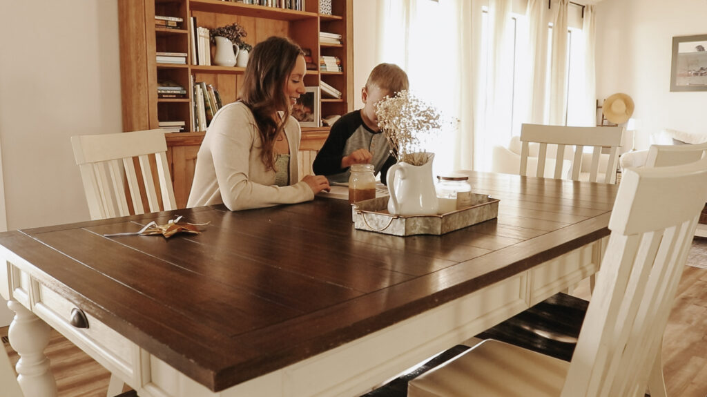Christian homemaker and mother sits at wooden dining table with child and teaches.