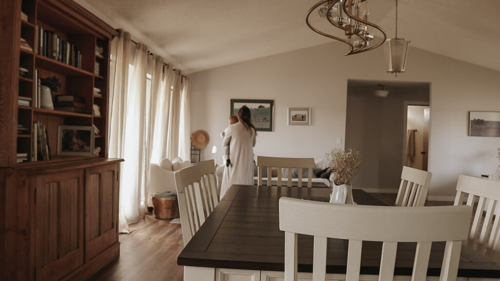 Christian homemaker walks to sofa while holding her baby in a bright, airy living room.