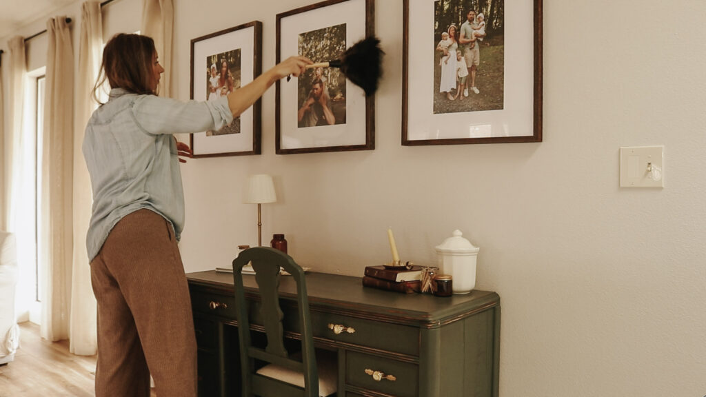 Homemaker dusts pictures hangin on wall above green desk.