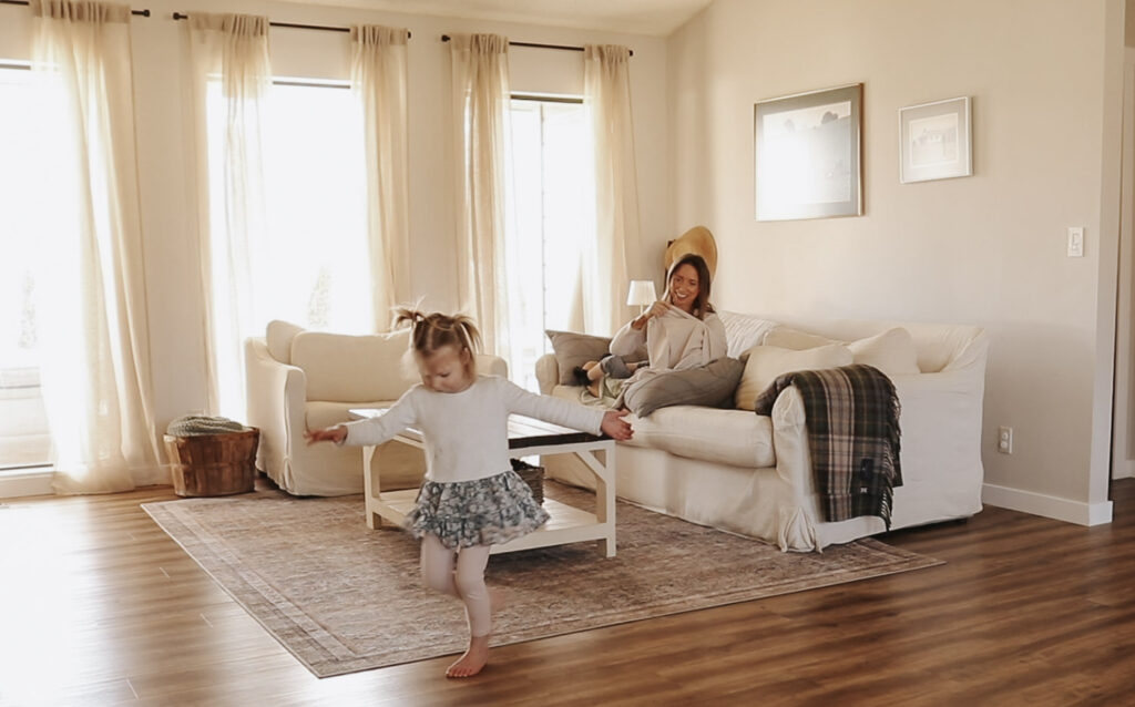 Homemaker and mother smiles while sitting on sofa holding baby and looking at toddler.