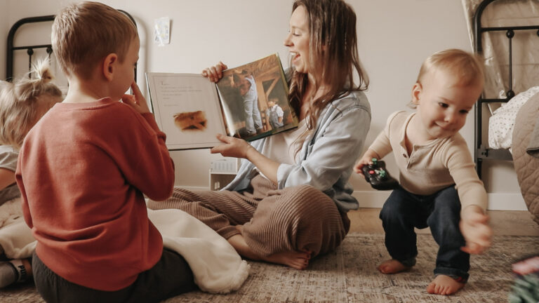Christian mother and homemaker sits on floor with children and reads a book while smiling.