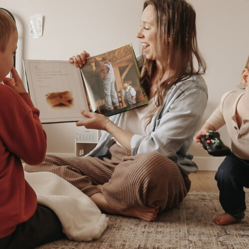 Christian mother and homemaker sits on floor with children and reads a book while smiling.